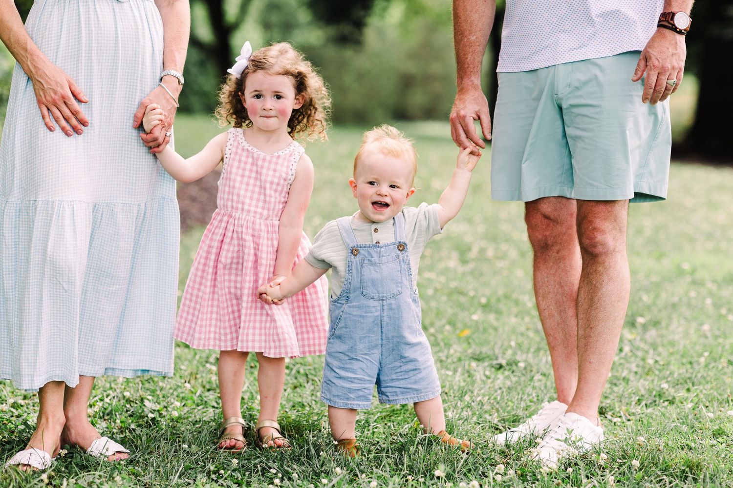 Two young siblings, a girl in pink and a toddler boy in overalls, hold hands outdoors.