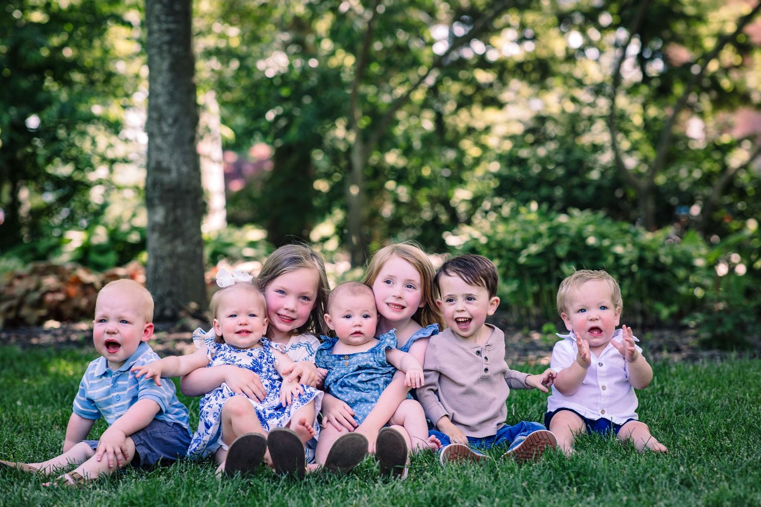 Seven happy babies and toddlers sitting together on green grass outdoors surrounded by lush trees.