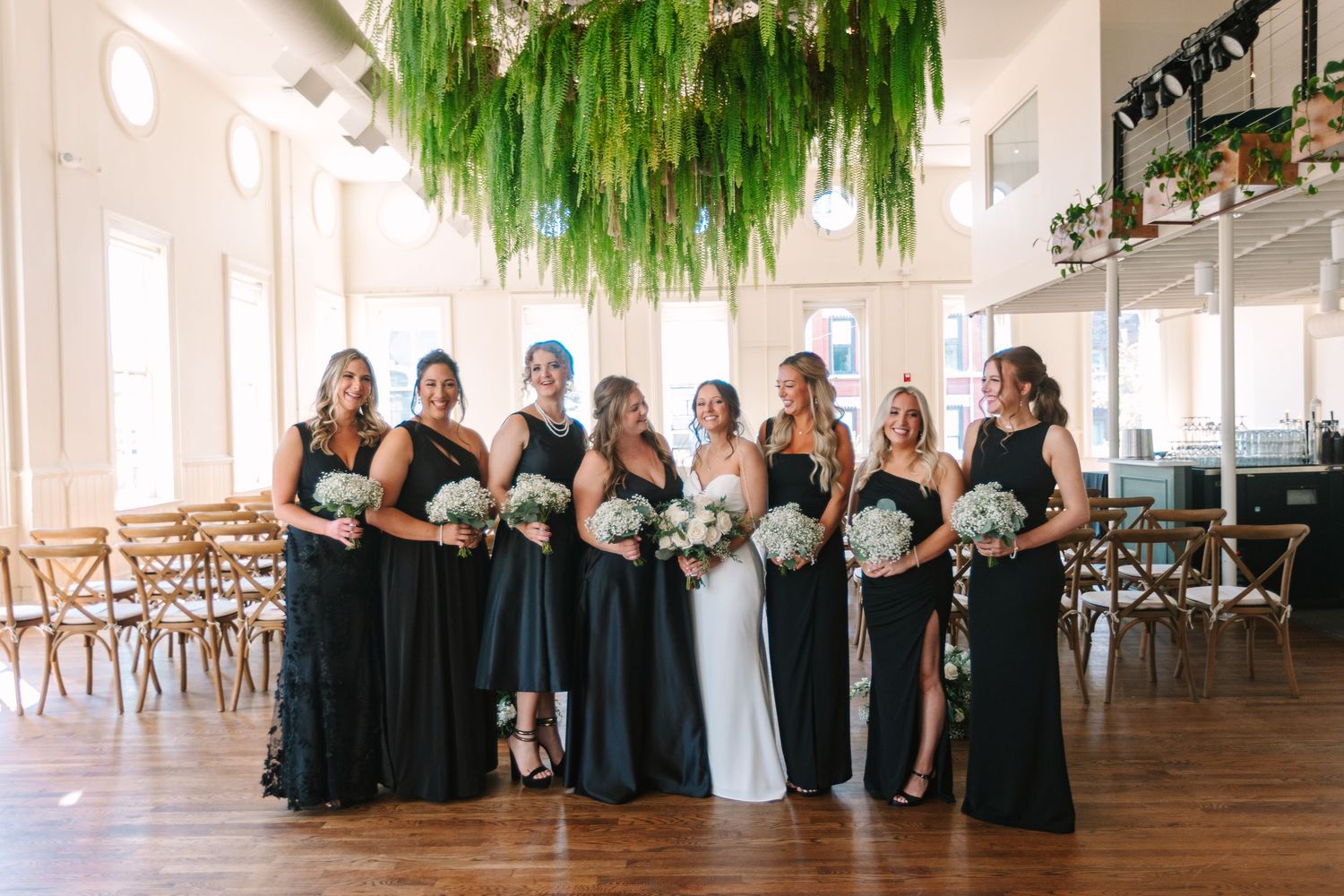 Bride and bridesmaids in black gowns posing in a bright venue with lush green hanging decor at the alcove by madtree.