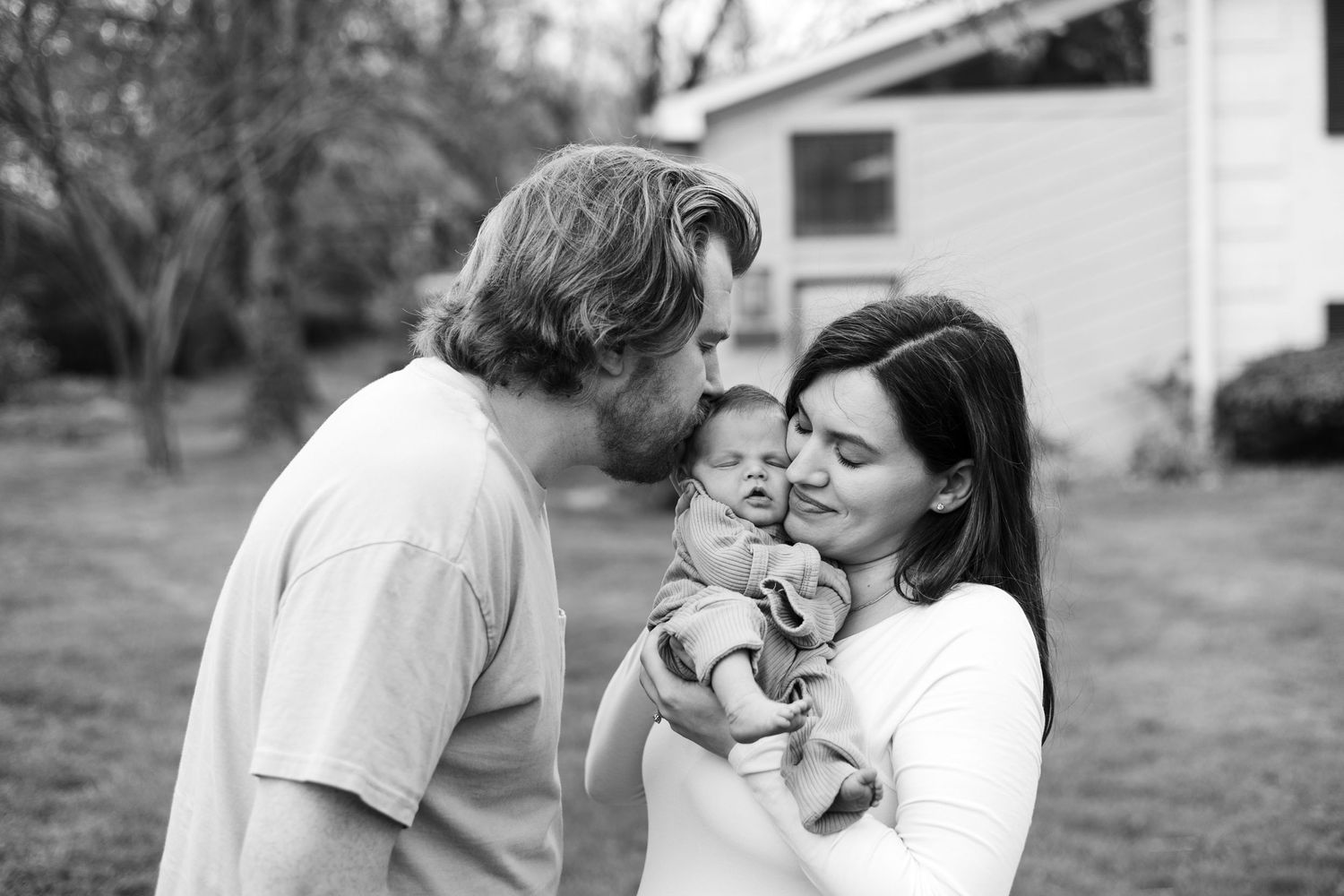 Black and white photo of parents kissing their newborn baby outdoors in a backyard.