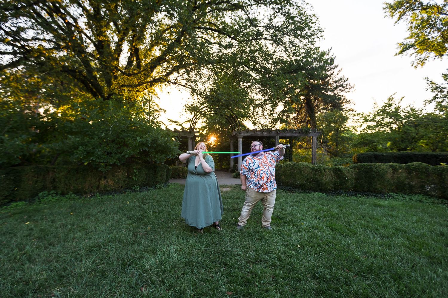 Couple playing with light sabres on a green lawn at sunset in a park.