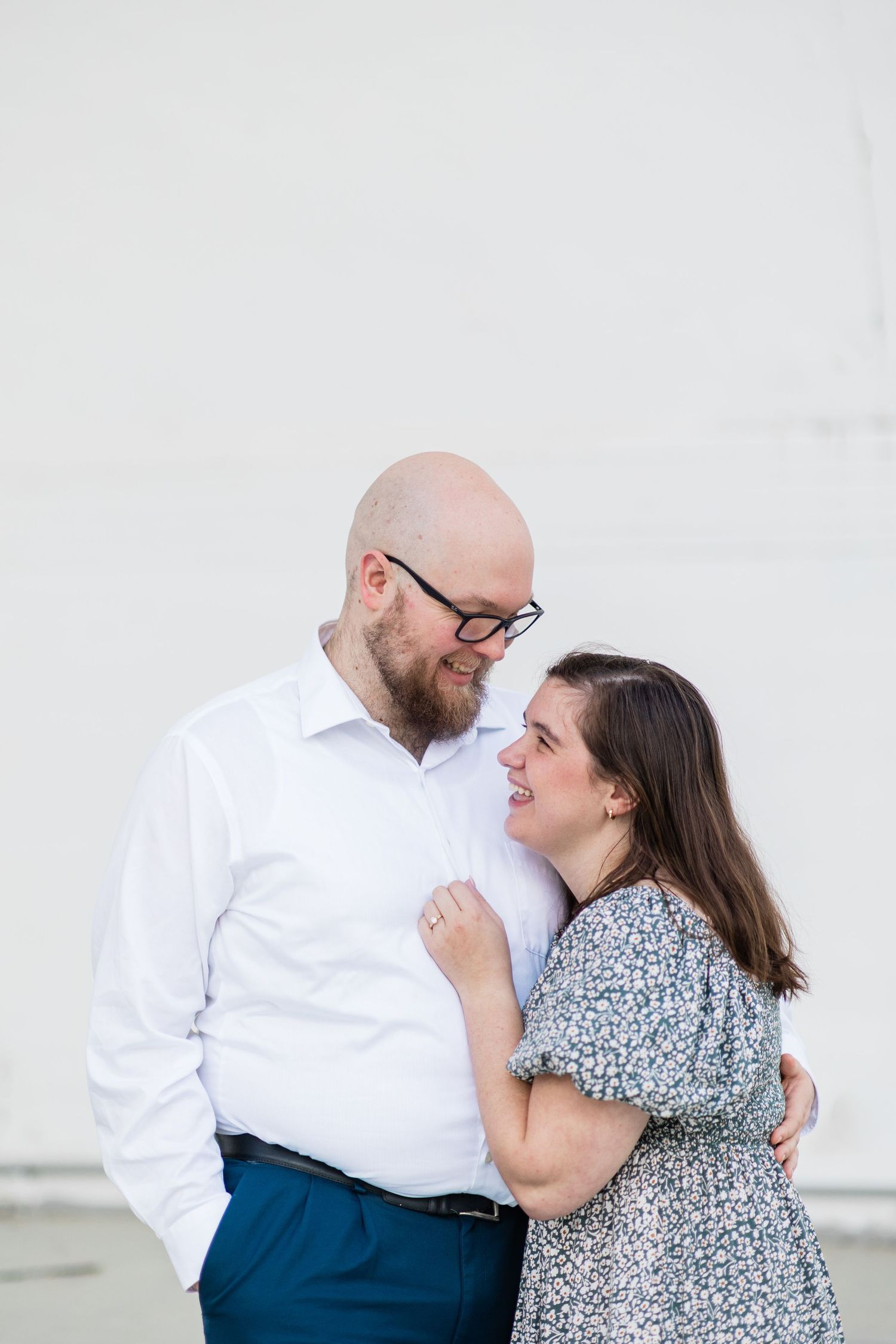 A bald bearded man in a white shirt embraces a brunette woman in a floral dress against a white wall.