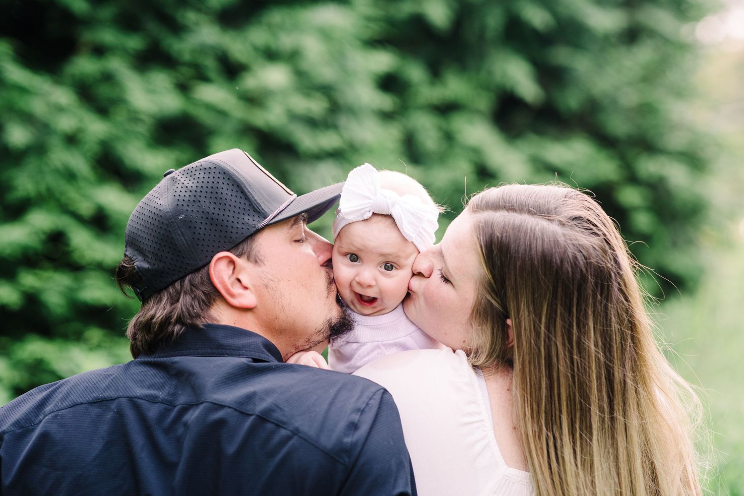 Two parents lovingly kiss their smiling baby girl on the cheeks outdoors in a lush green setting.
