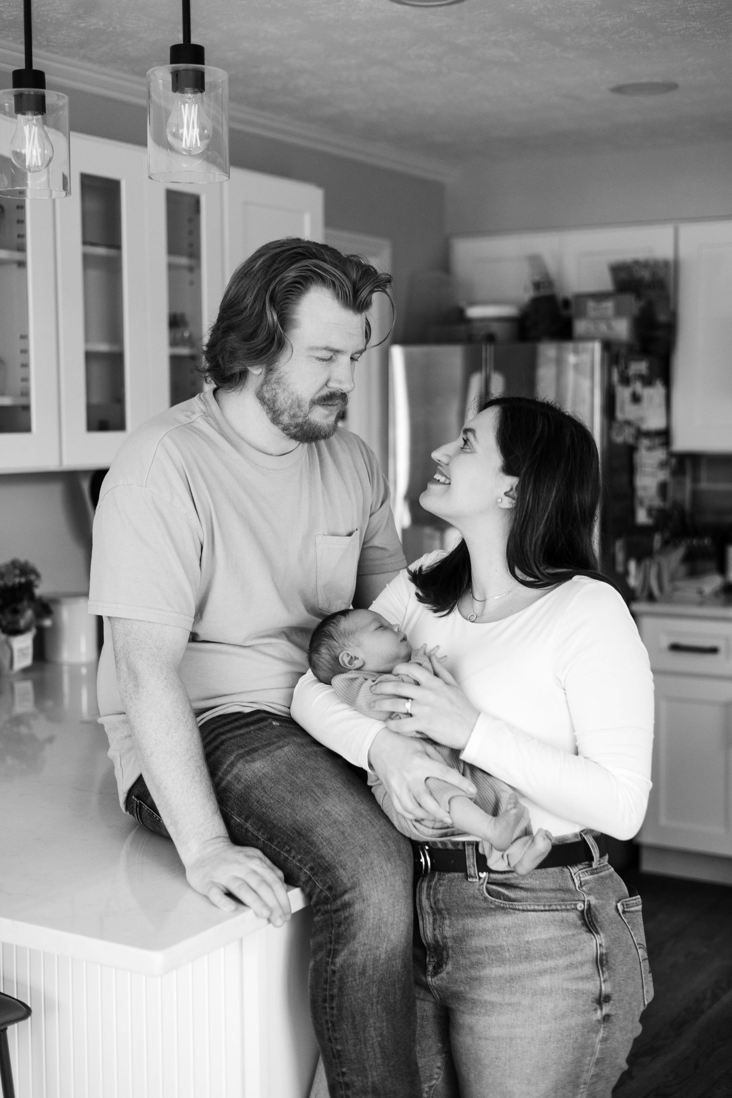 Black and white photo of a couple smiling at each other in a kitchen while holding their newborn baby.