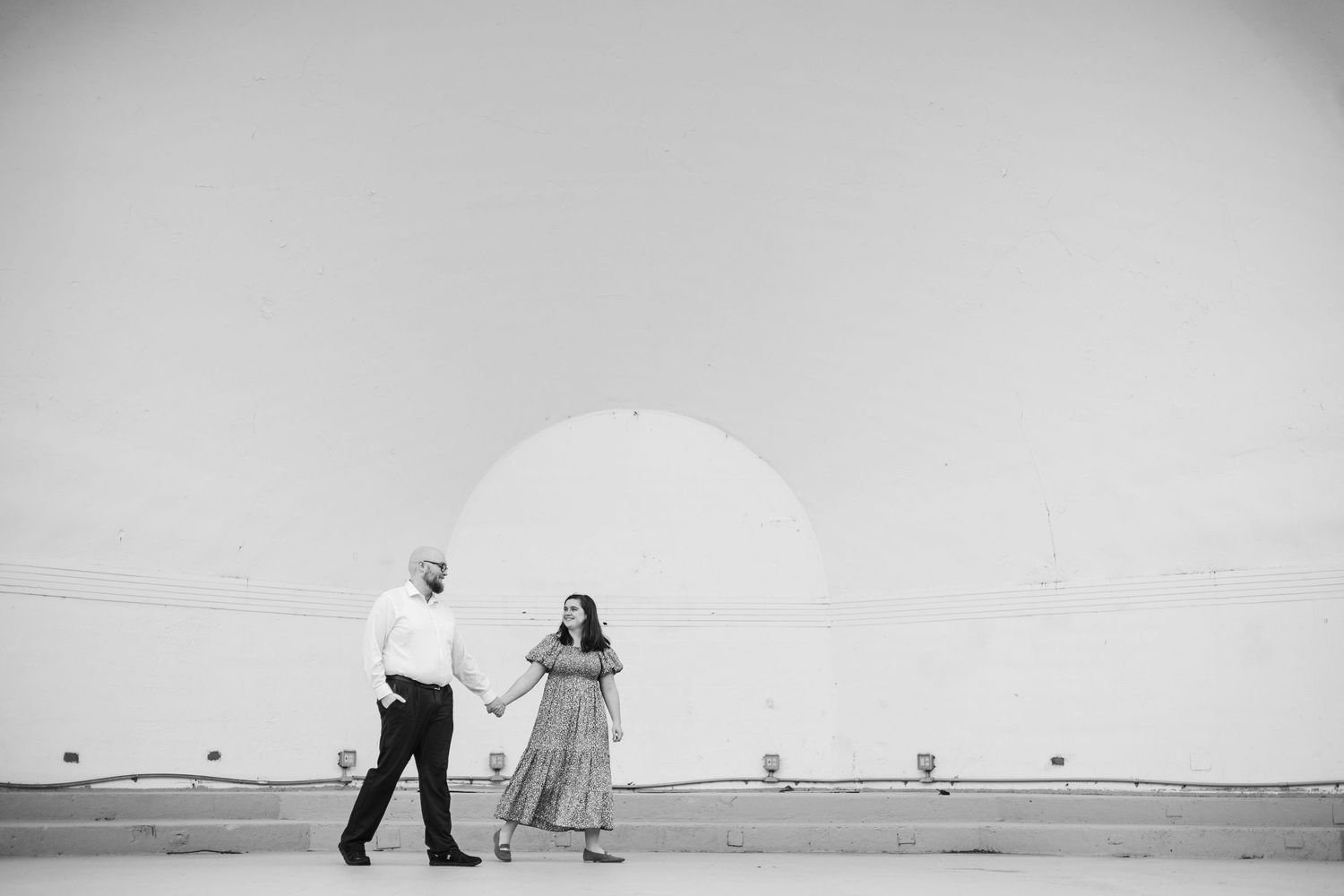 Black and white photo of an engaged couple holding hands walking outdoors with large industrial structures behind them.
