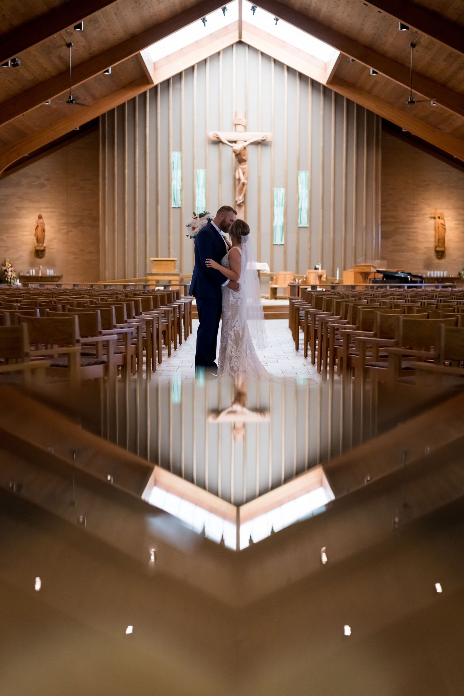 Bride and groom kissing at church altar reflected in polished floor with crucifix in background.
