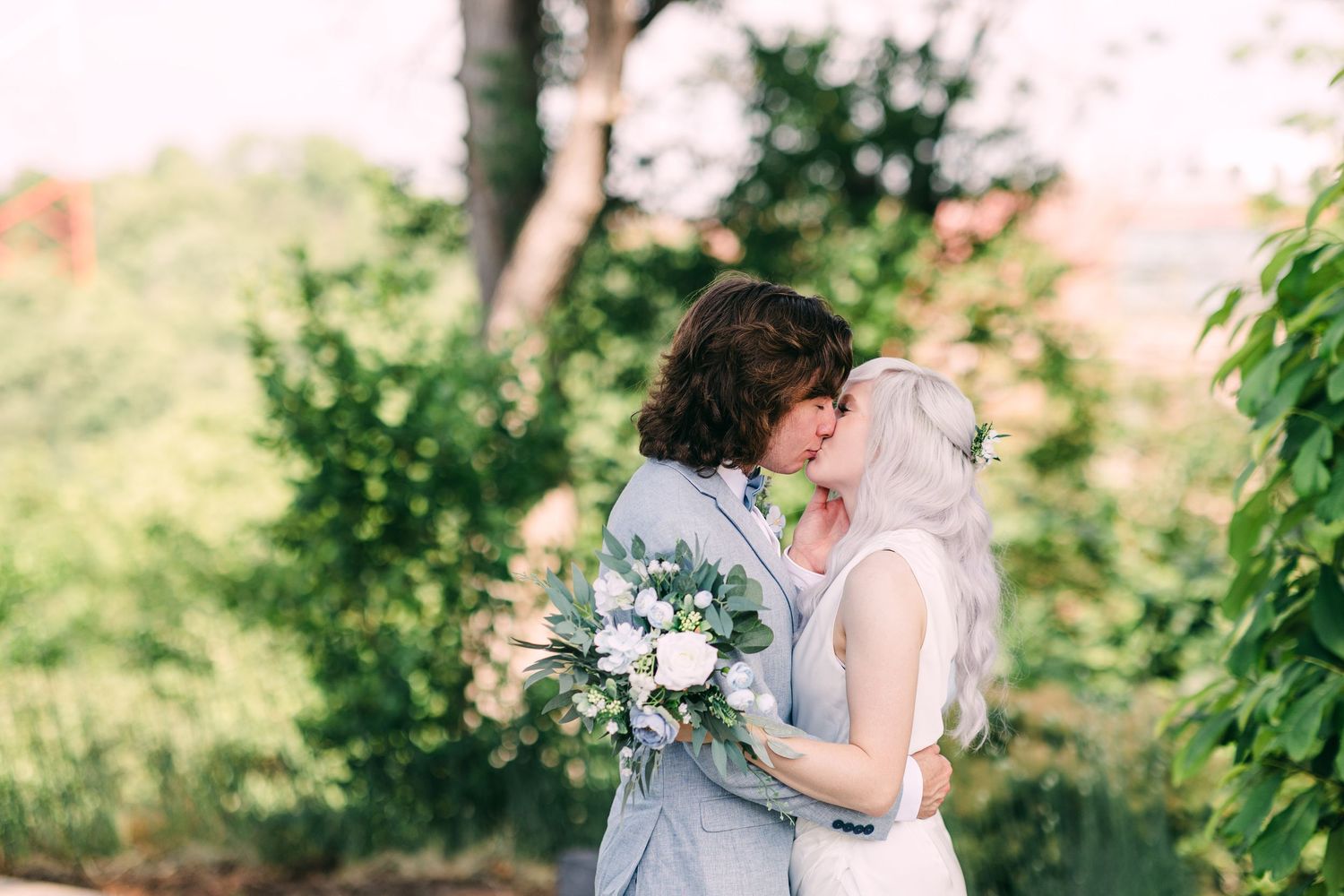 Bride with silver hair and groom in light blue suit share a tender kiss surrounded by lush garden greenery.