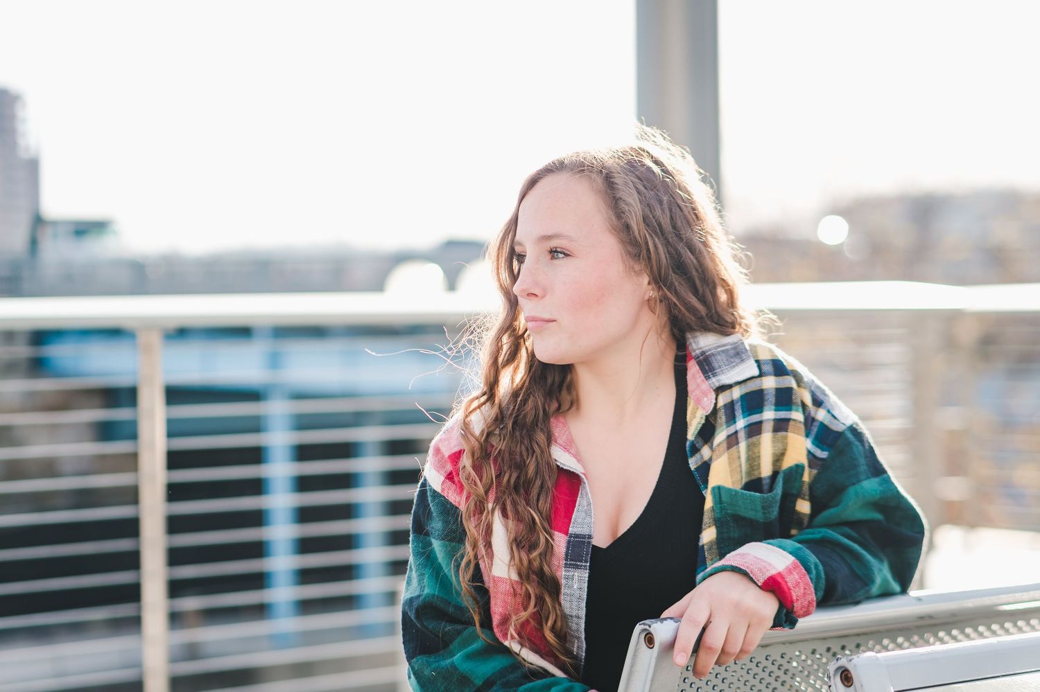 Young woman in colorful plaid jacket leaning on metal railing with bright sky background.