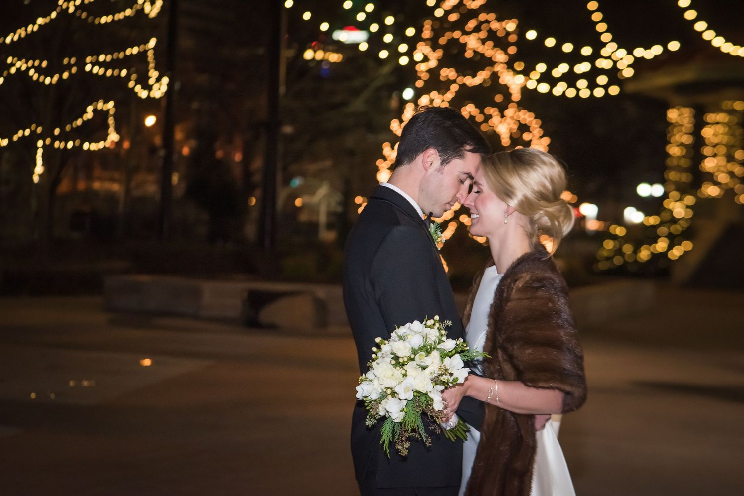 Romantic bride and groom sharing intimate moment outdoors at night with warm bokeh string lights behind them.