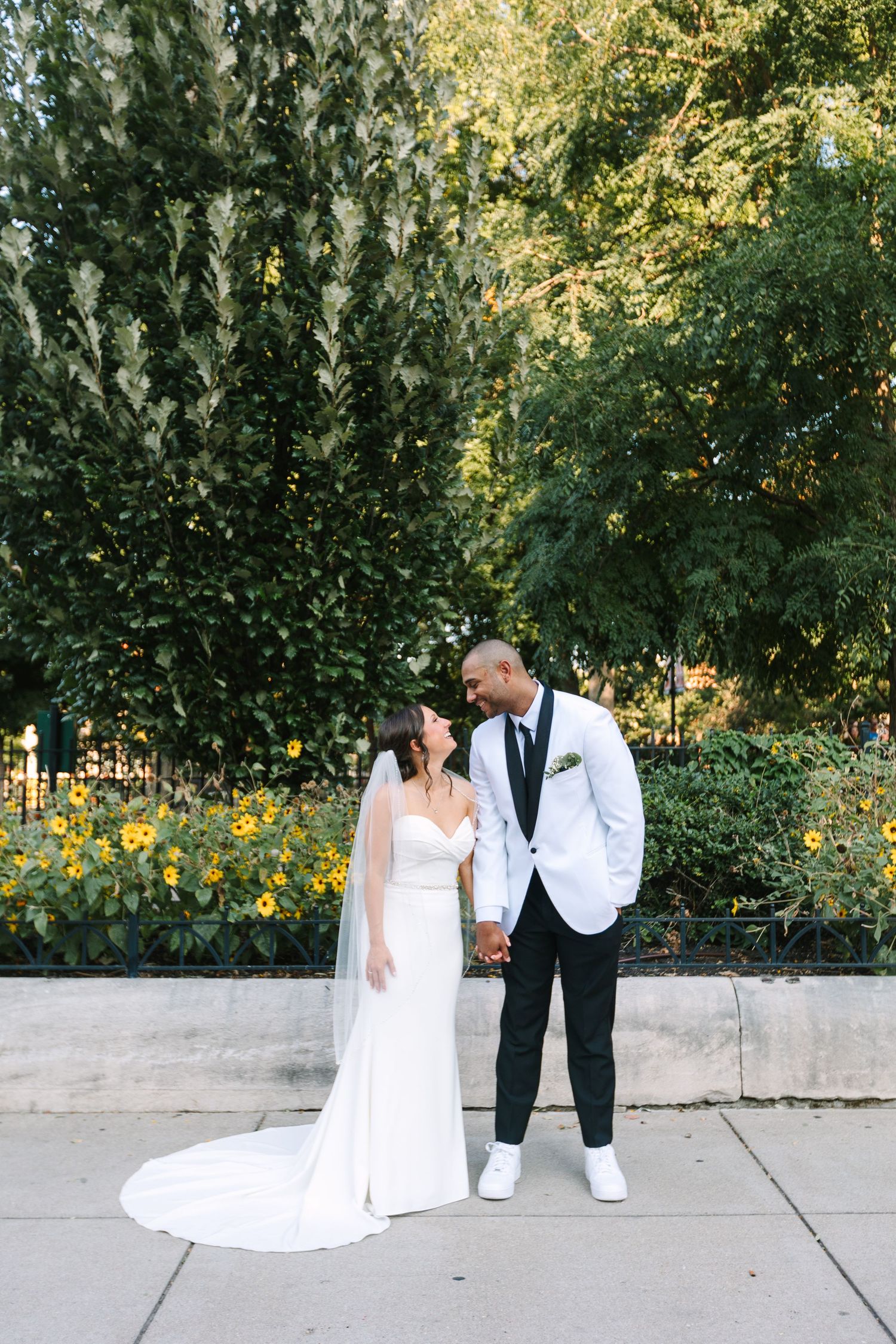 Bride and groom in elegant attire hold hands and smile outdoors among lush green trees.