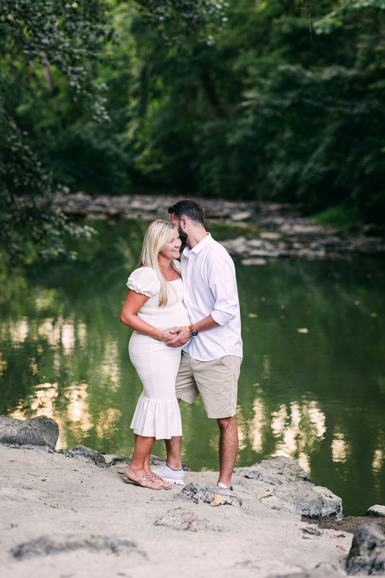 Pregnant couple embracing by a serene green river, woman in white dress, man in light blue shirt and khaki shorts.