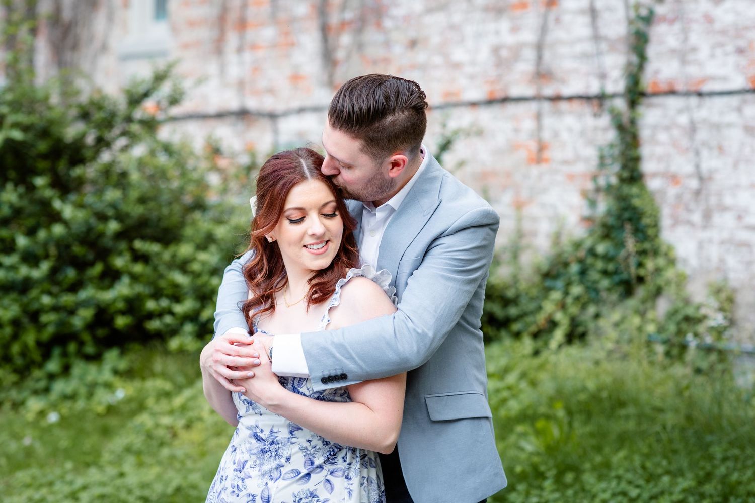 Man in grey suit embracing woman in floral dress from behind near a rustic stone wall in a garden.