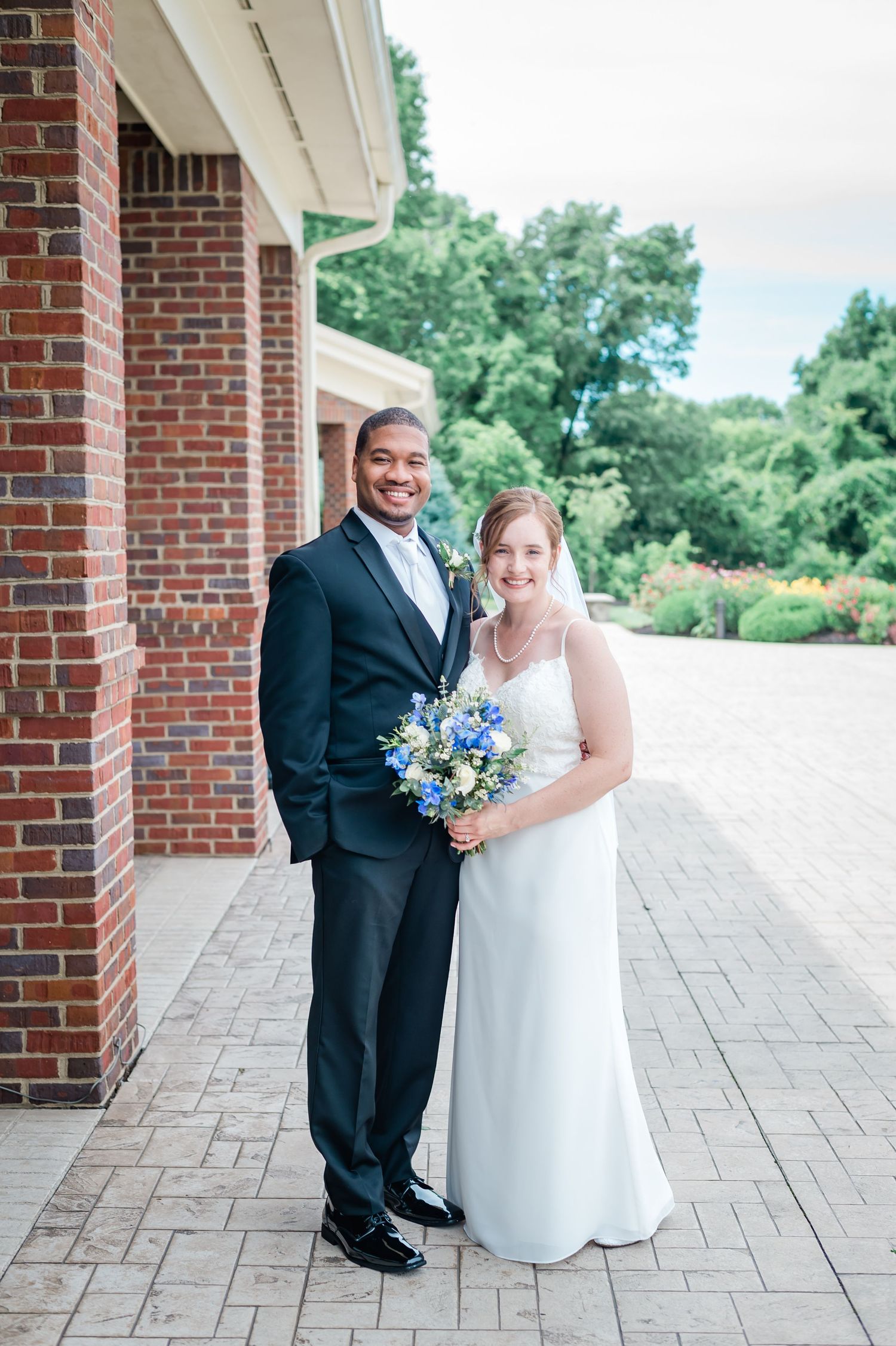Newlywed couple posing outside brick building, bride holding blue and white bouquet in white gown.