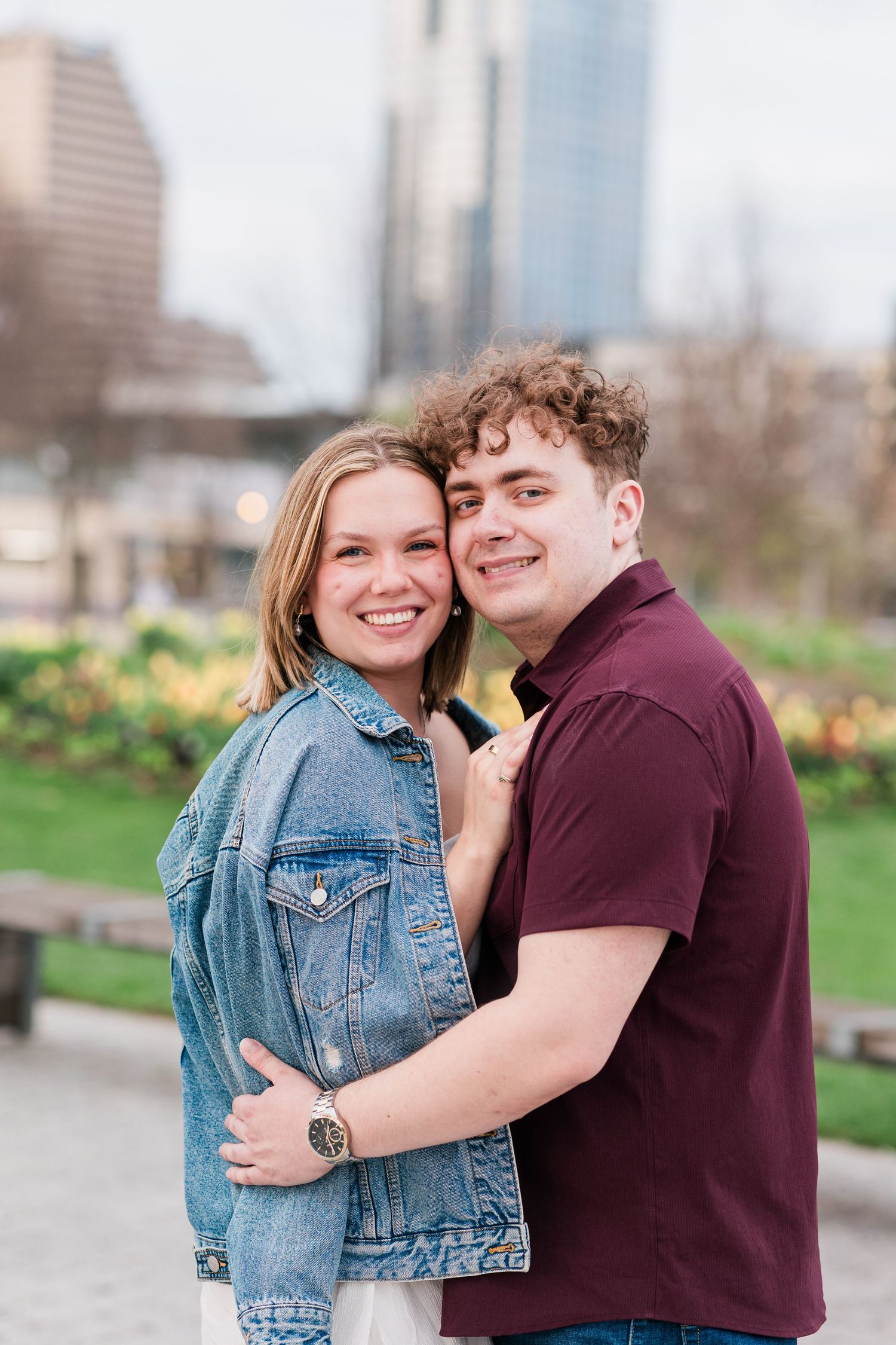 A smiling couple embraces and poses together in an outdoor urban park setting with tall buildings in the background.