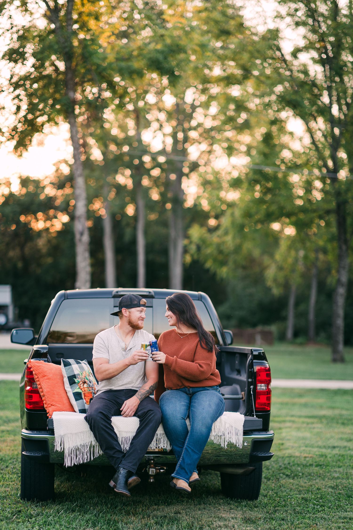 A couple sits on a truck tailgate toasting drinks and smiling at each other during a sunset session.