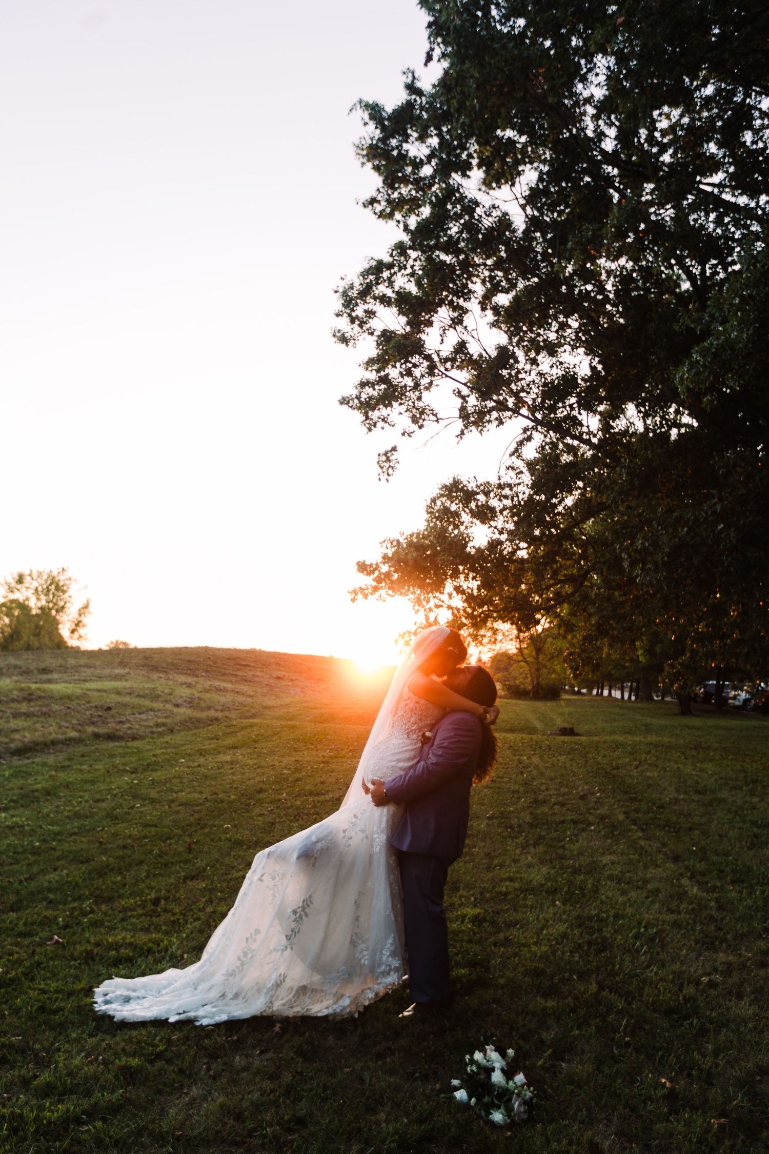 A bride and groom embrace at sunset on a lush green hill, with golden light glowing behind them near a large tree.