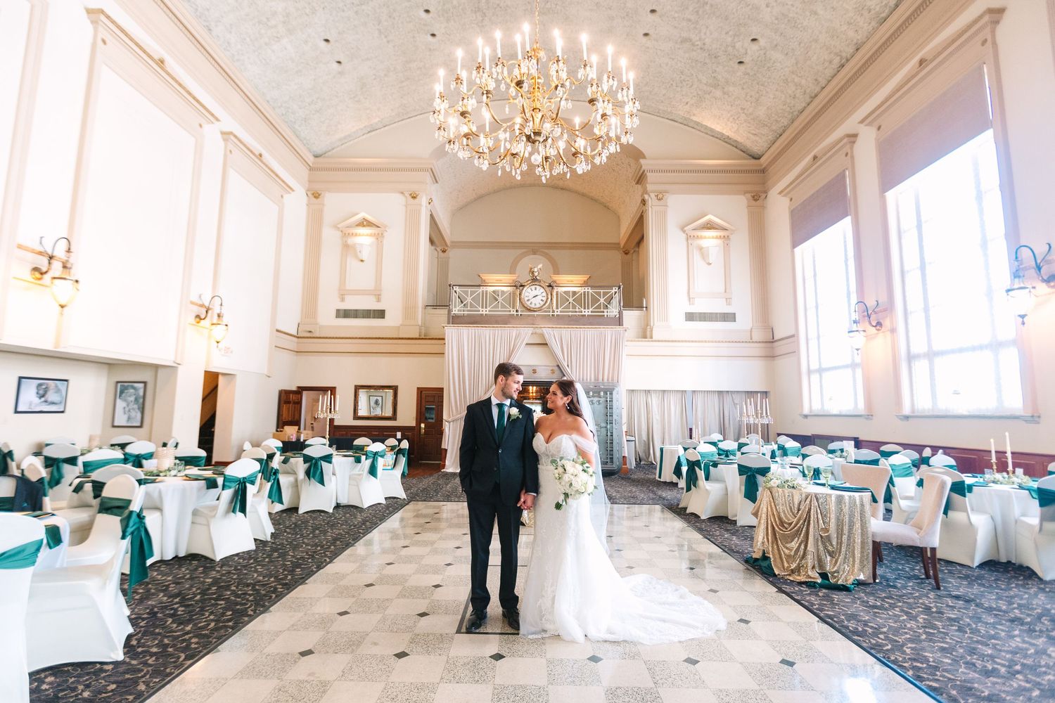 Bride and groom standing in grand ballroom with crystal chandelier, teal chair sashes, and elegant decor.