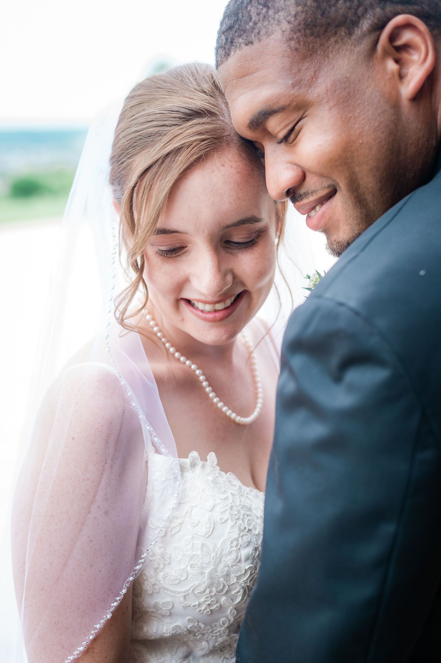 Close-up of smiling bride and groom touching foreheads, bride wearing pearl necklace and veil.