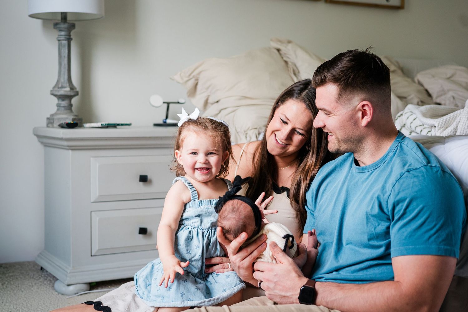 Happy young family with toddler daughter smiling together on bedroom floor with newborn baby.