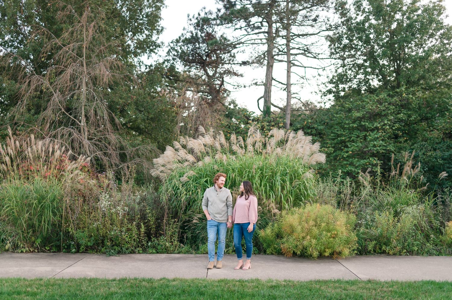 Couple holds hands standing on pathway in front of lush ornamental grasses and mature trees.