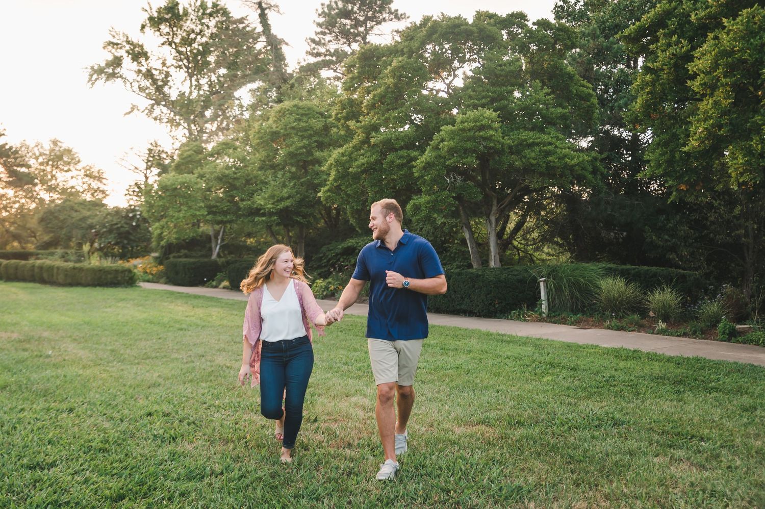 Happy couple holds hands walking through lush green park lawn during golden hour sunset.