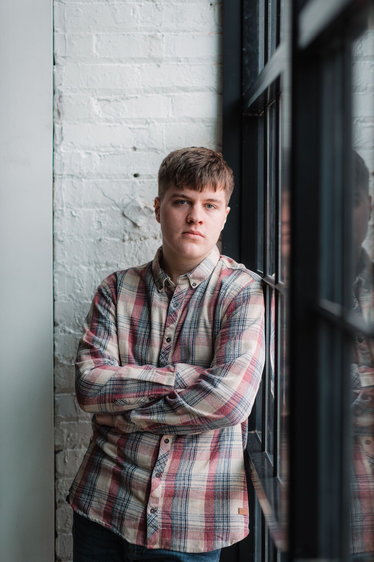 Young man in plaid shirt stands with arms crossed leaning against white brick wall by window.