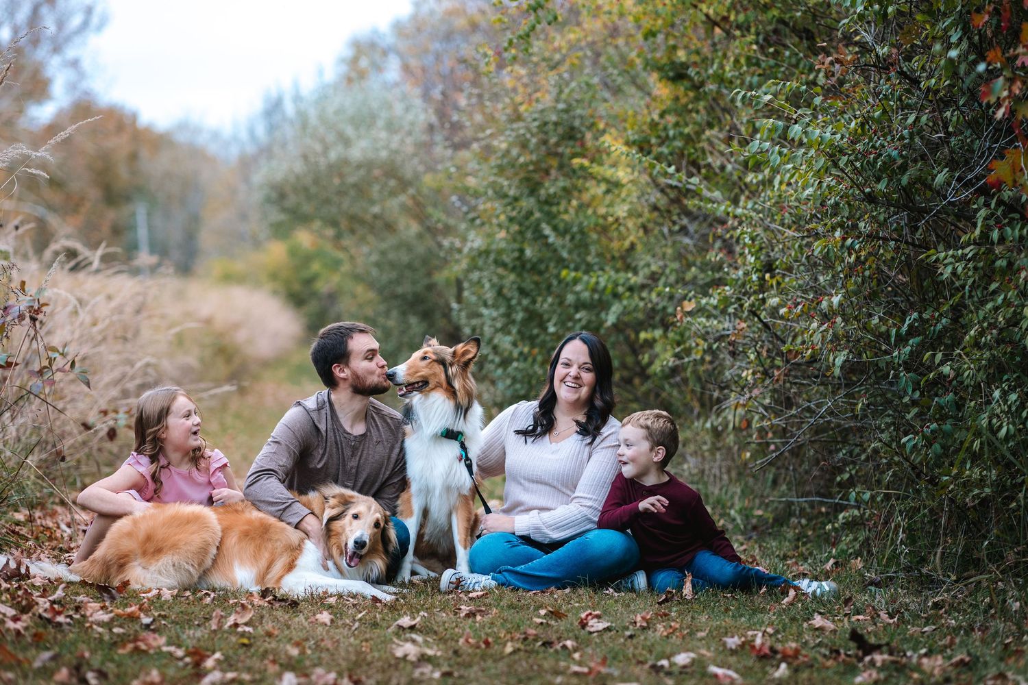 Happy family sitting outdoors with two Collie dogs among fall foliage.