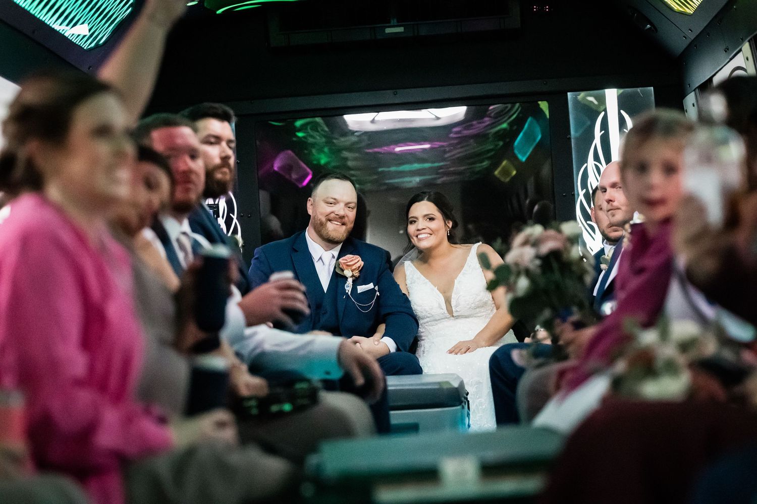 Bride and groom smile joyfully inside a decorated party bus surrounded by wedding guests.