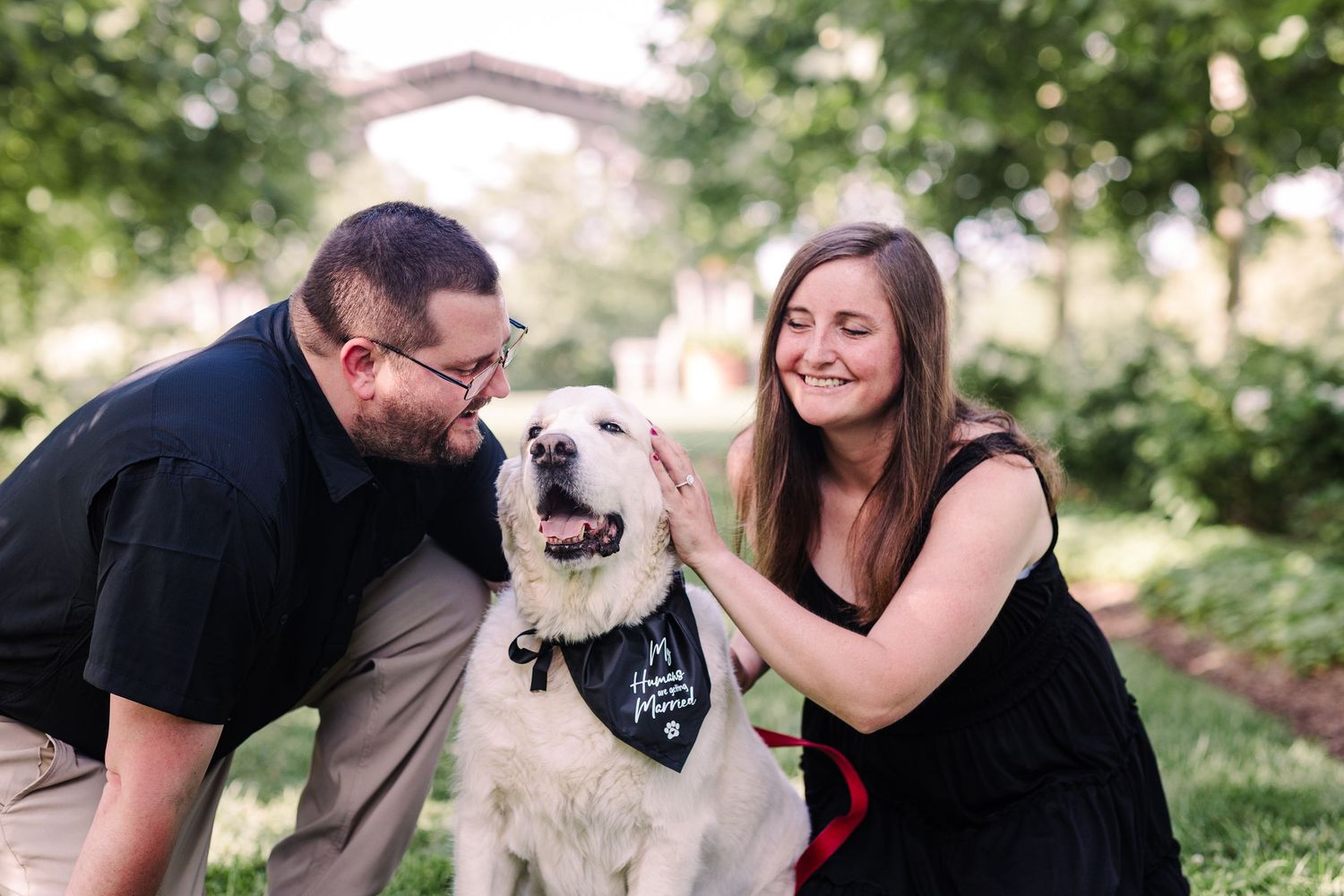 A smiling couple pets their white Golden Retriever wearing a wedding bandana during an engagement photo session.