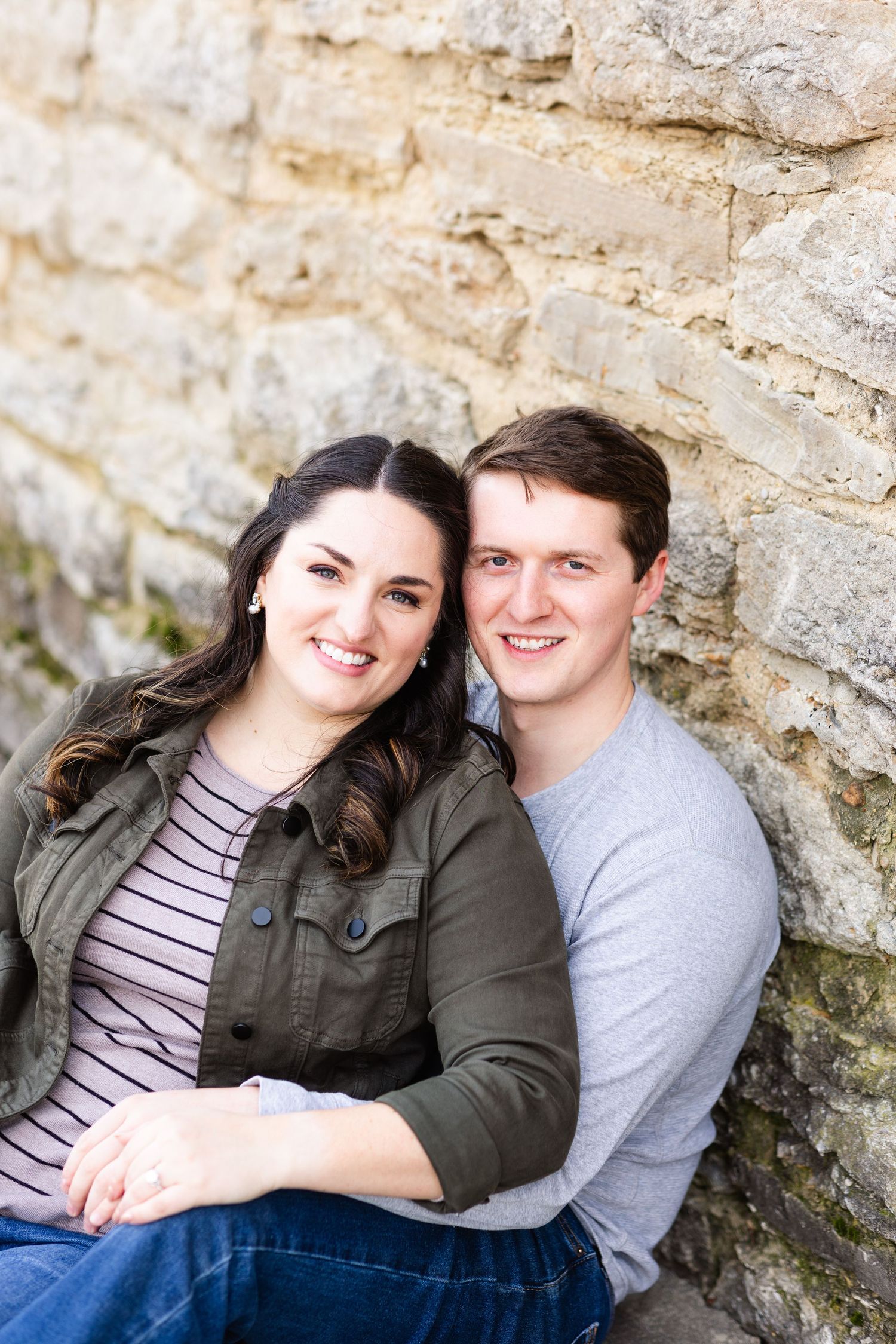 Smiling couple sitting together against a rustic stone wall outdoors.