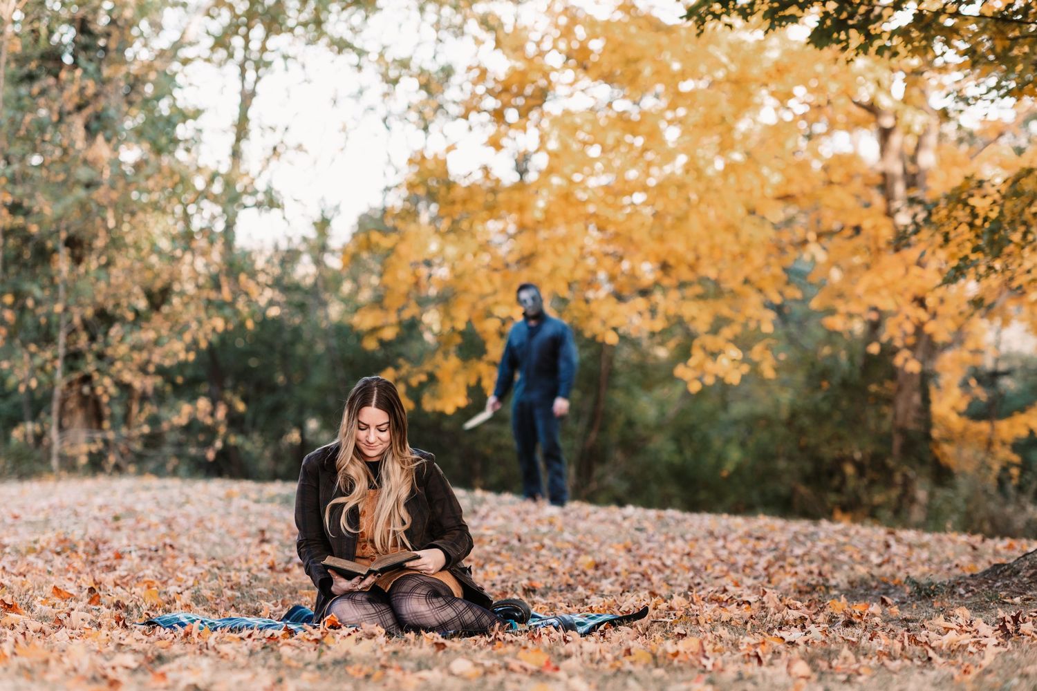 Woman sitting in autumn leaves reading while man stands in background among fall trees.