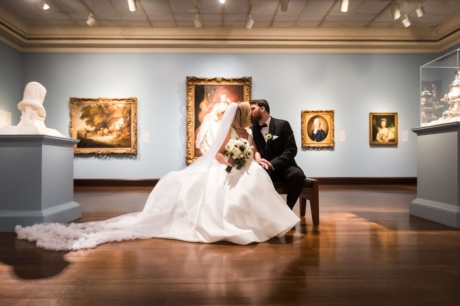 Bride and groom share romantic moment seated in Cincinnati art museum gallery surrounded by classic paintings.