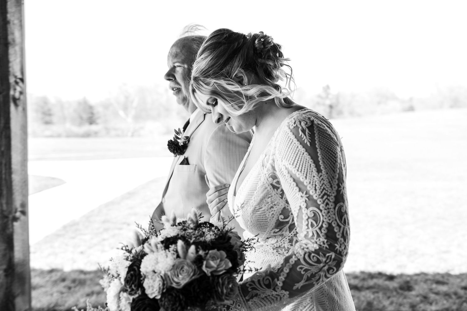 Emotional bride in lace wedding gown holds bouquet in black and white wedding portrait as she walks with her father.