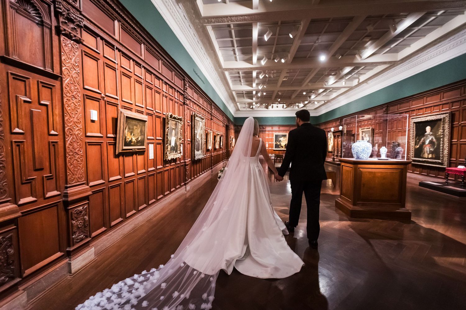 Bride and groom walk hand in hand through grand wood-paneled Cincinnati art museum gallery corridor.