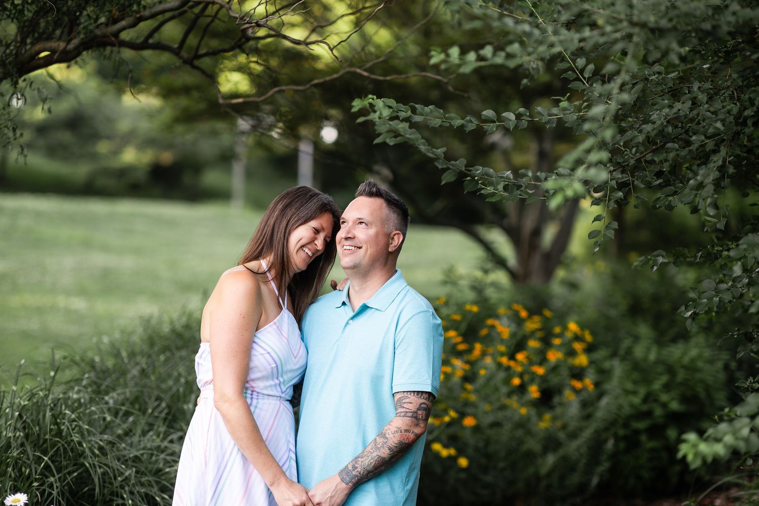 Smiling couple standing close together under a large tree in a lush green park during golden hour.