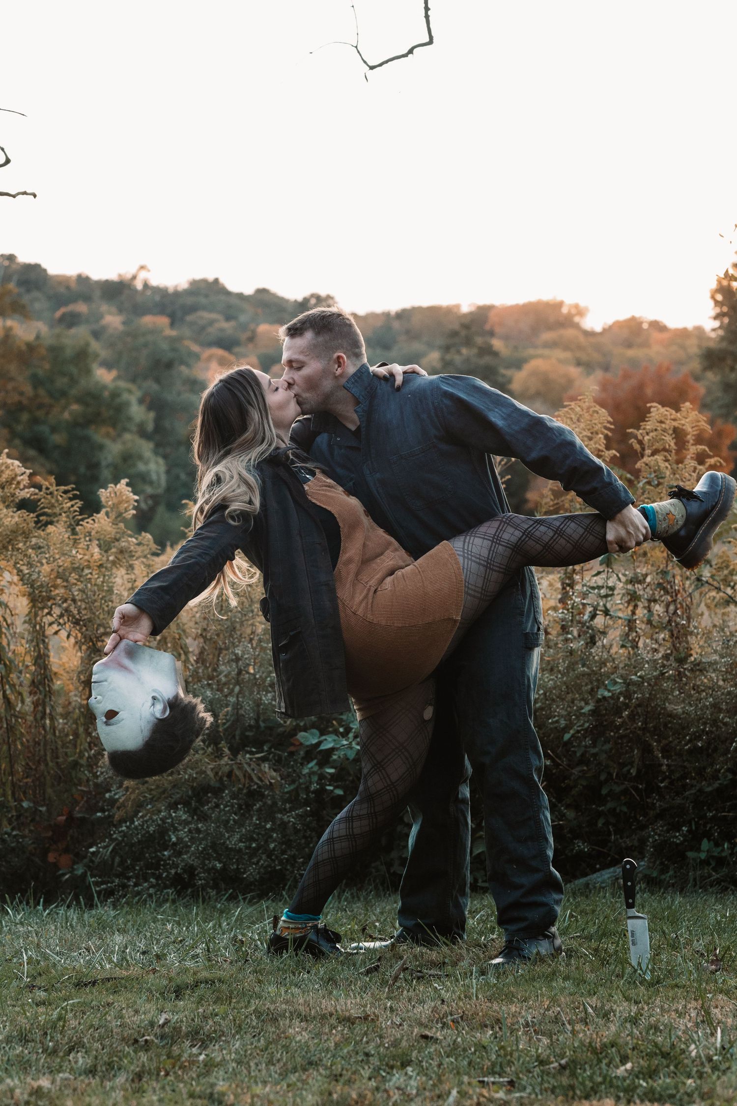 Man in Halloween costume removes Michael Myers mask while romantically dipping and kissing woman outdoors in fall setting.