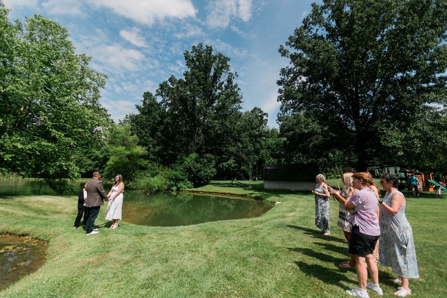Small outdoor wedding ceremony beside a serene pond surrounded by lush green trees with guests gathered on the lawn.