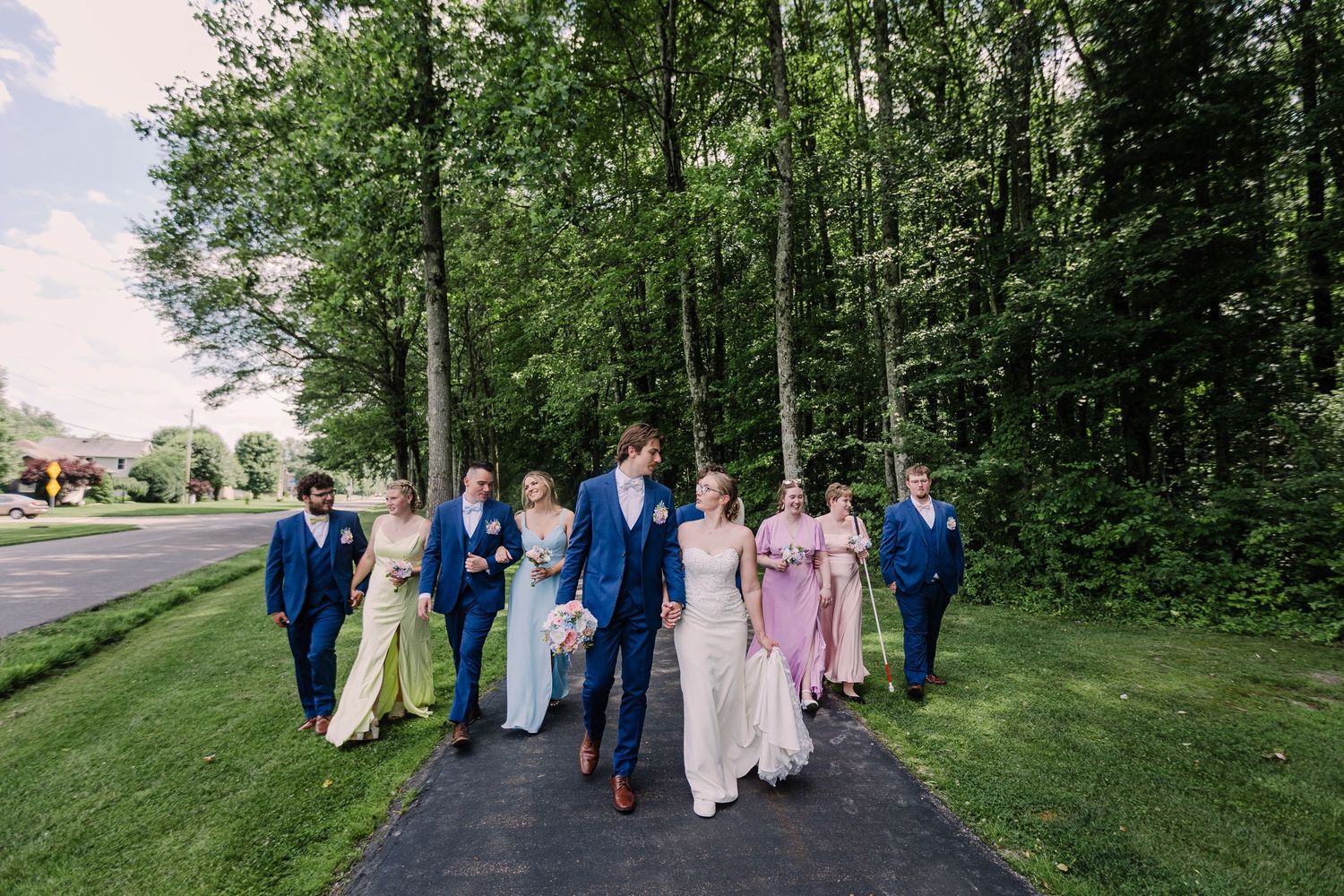 Wedding party walking down a tree-lined path in colorful formal attire on a sunny day.
