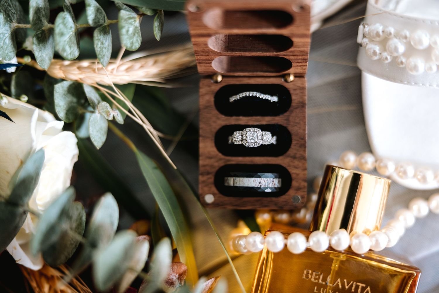 Wedding flat lay featuring diamond rings in a wooden box, pearl jewelry, and Bellavita perfume.