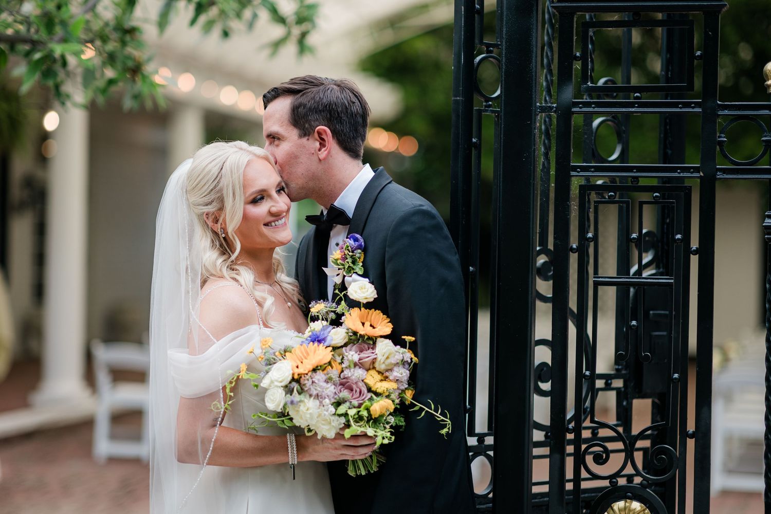 Smiling bride holding colorful bouquet receives a tender kiss from groom beside ornate black iron garden gate.