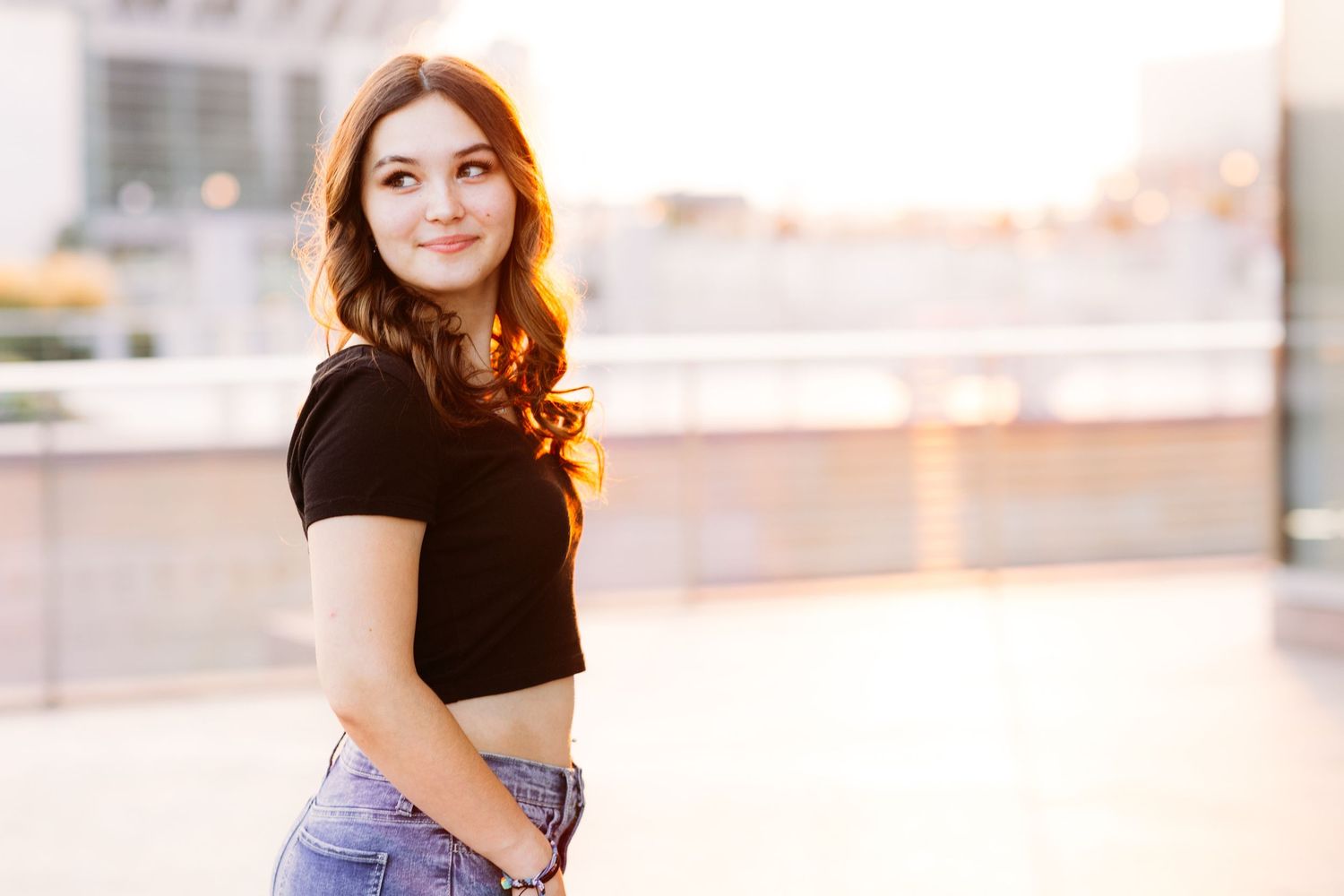 Young woman in black crop top and jeans smiling over her shoulder in golden sunset light.