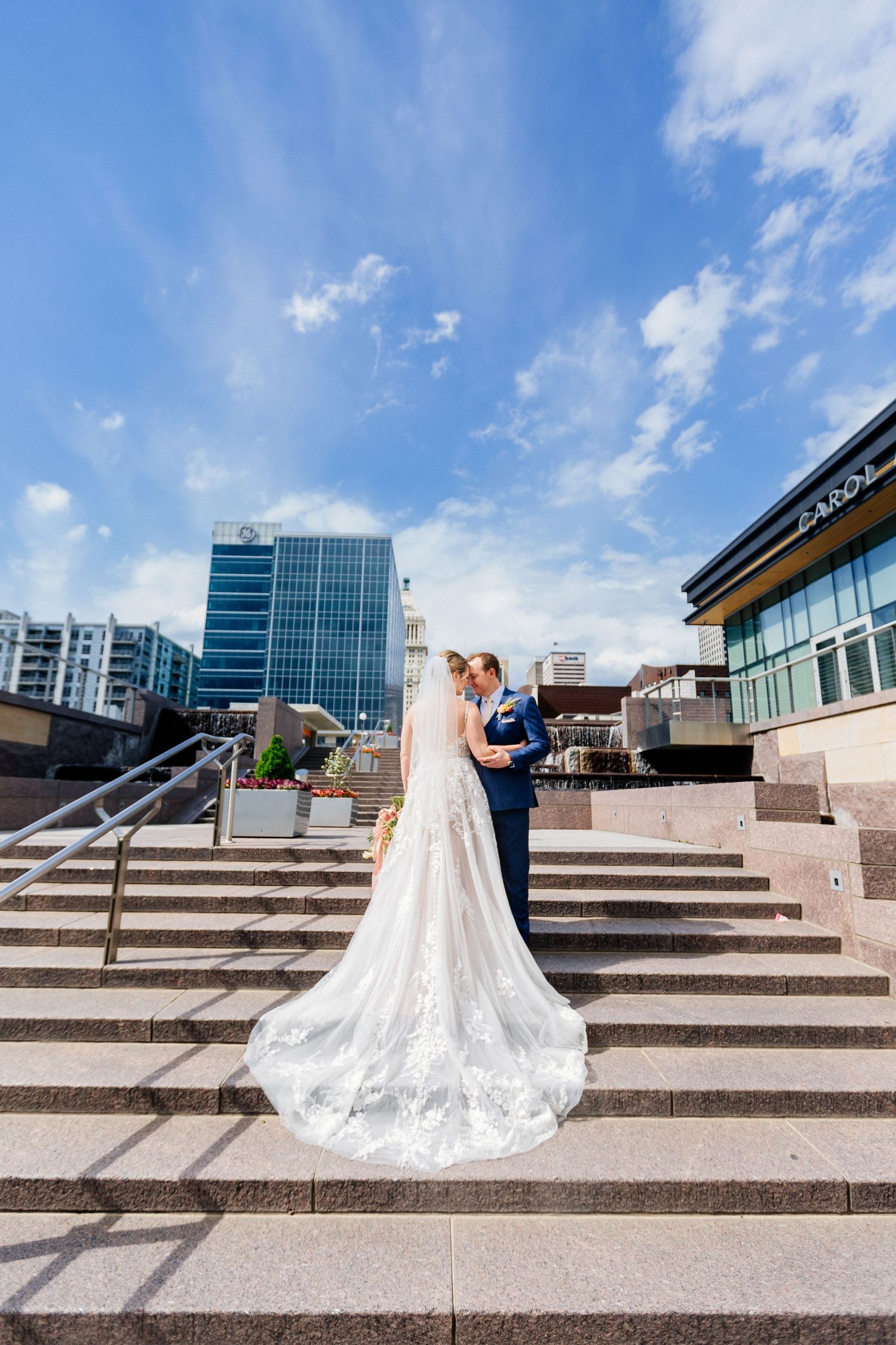 Bride and groom kissing on wide stone steps with a city skyline and blue sky in the background.