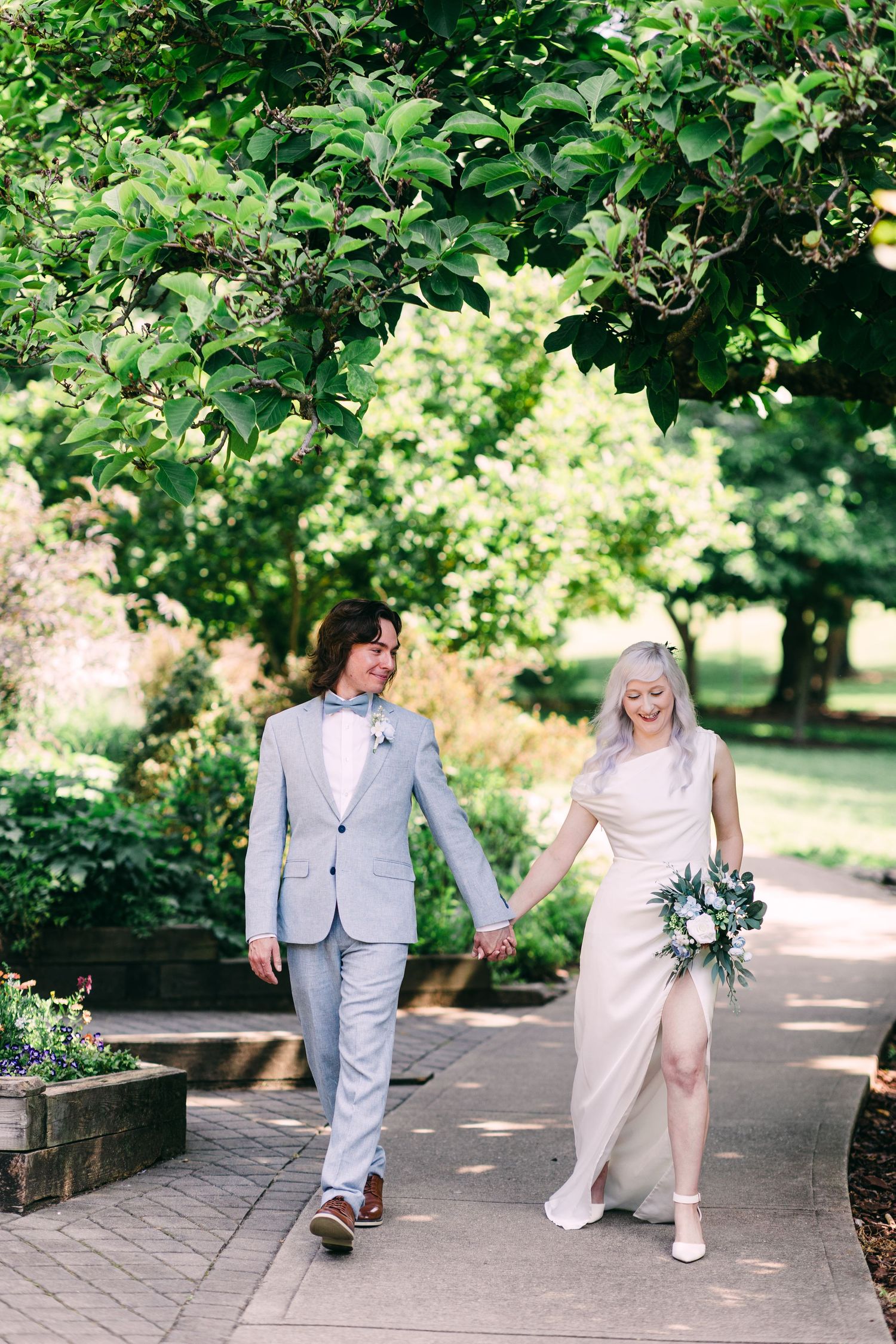 Newlywed couple walks hand in hand under lush green tree canopy, bride holding white bouquet in garden.