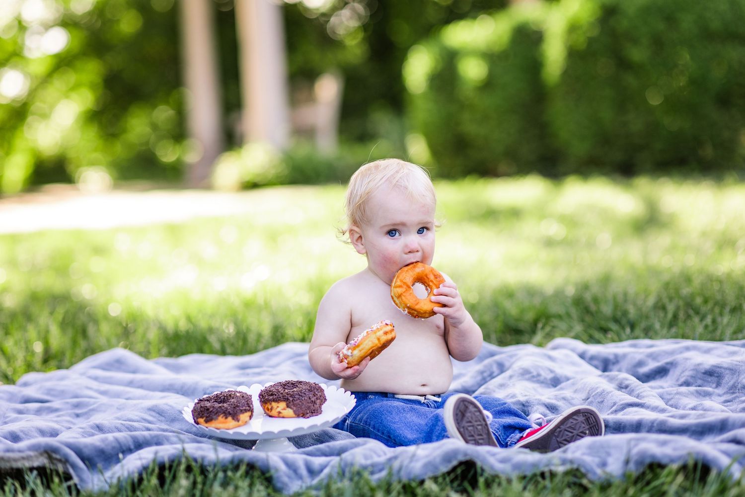 Shirtless baby boy sitting on blanket in park eating colorful donuts on a sunny day.