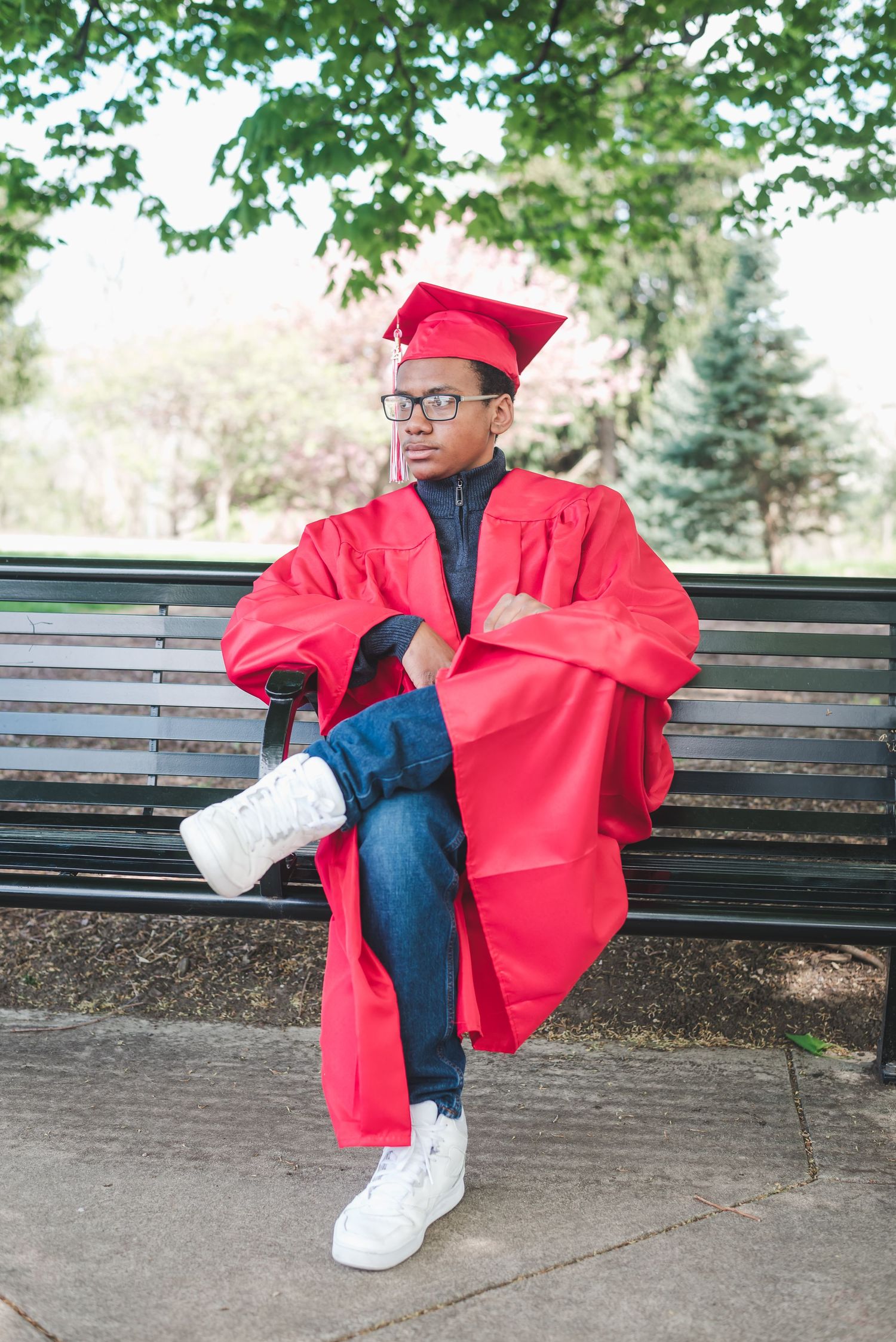 Graduate in red cap and gown sitting cross-legged on park bench with trees behind.