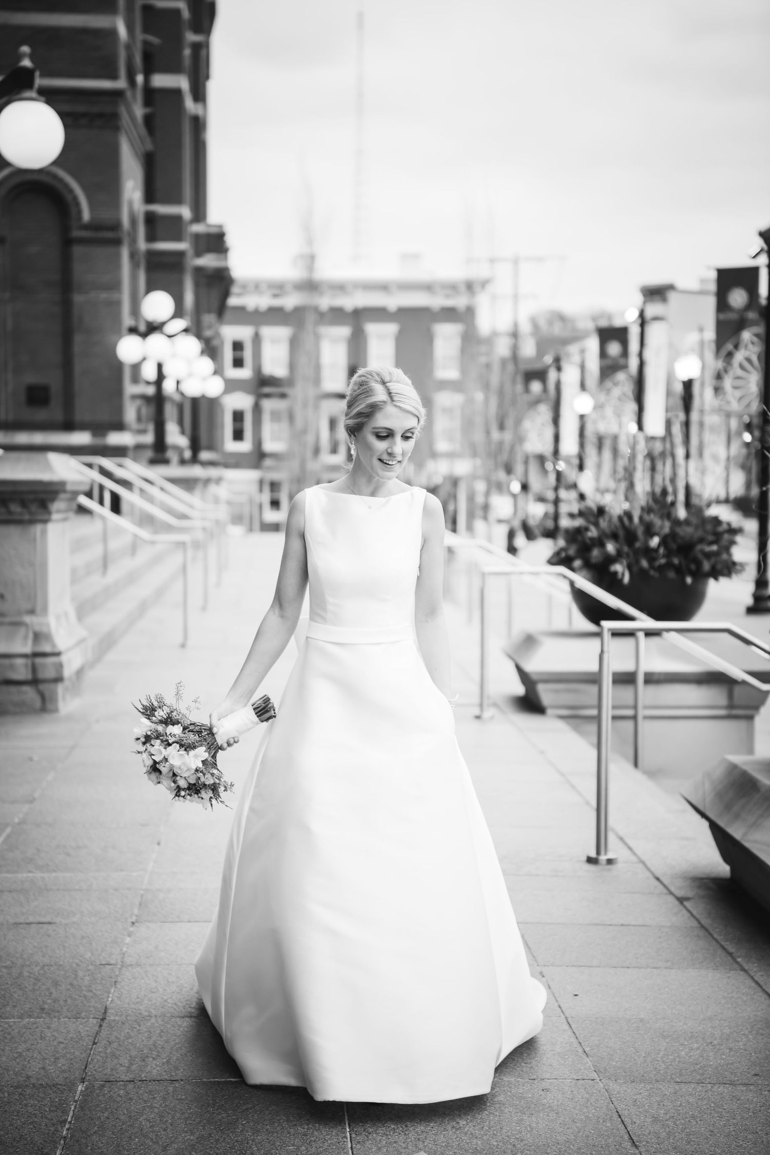 Elegant bride in classic white gown holding bouquet on urban plaza in black and white.