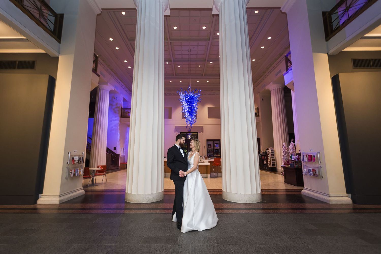 Bride and groom embrace between grand white columns in a beautifully lit Cincinnati Art Museum.