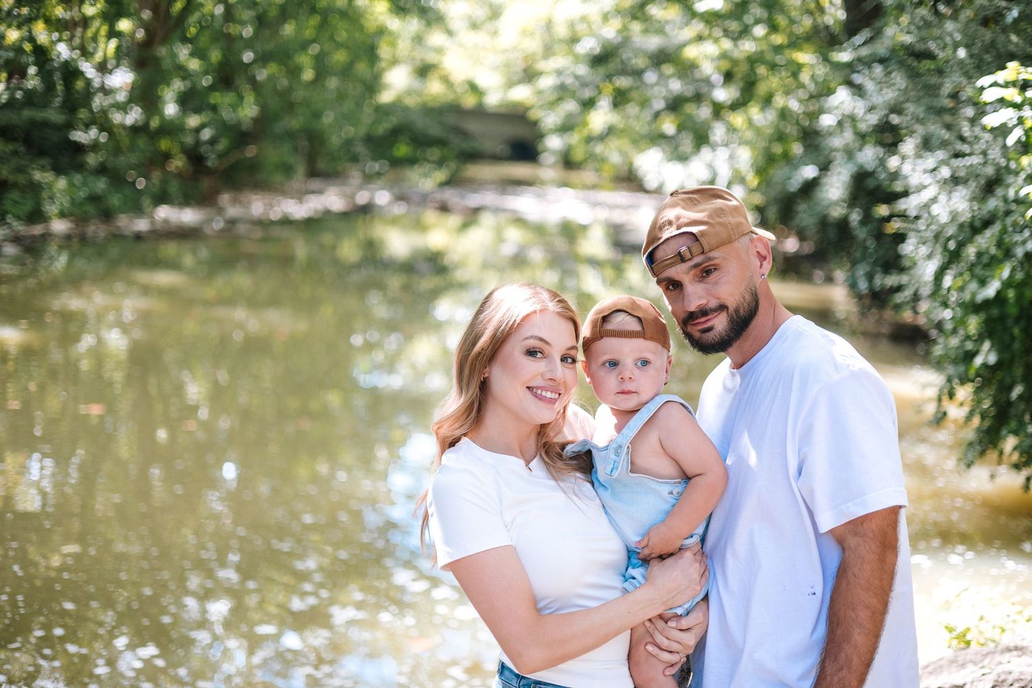 Young family of three smiling together near a serene green pond on a sunny summer day.