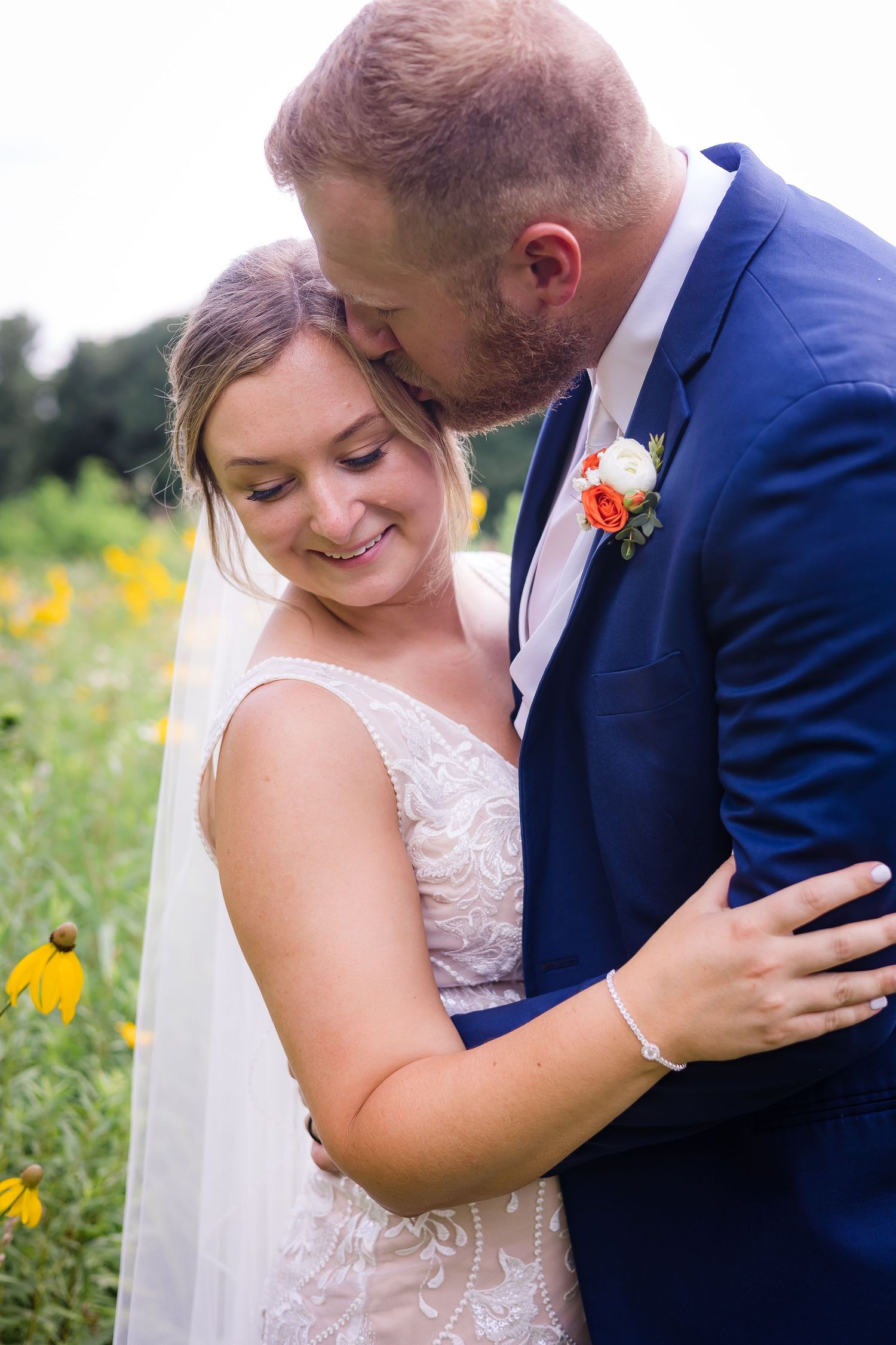 Bride and groom embracing in a wildflower field, groom in navy suit kissing brides temple as she smiles softly.