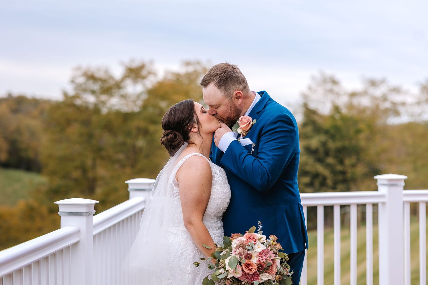 Bride and groom sharing a romantic kiss on a white deck with fall foliage.